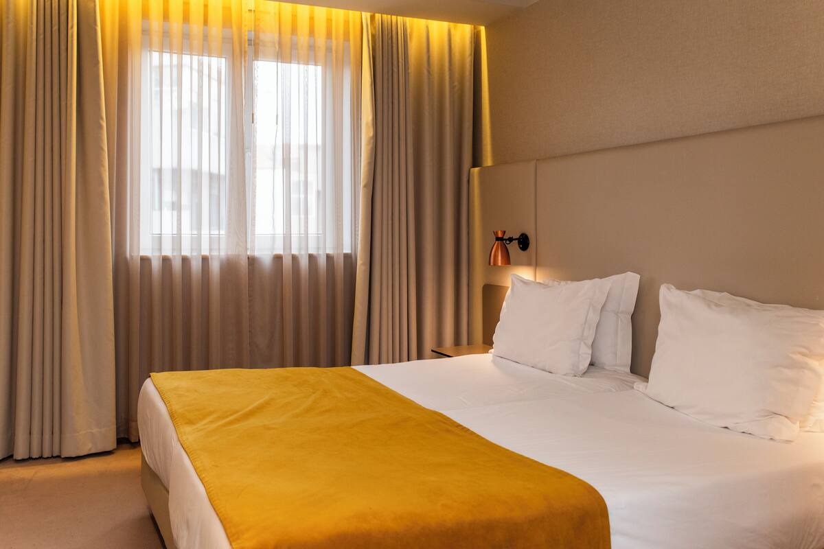 A hotel room with a large bed, beige curtains, a bedside lamp, and a view of the outdoors through the window.