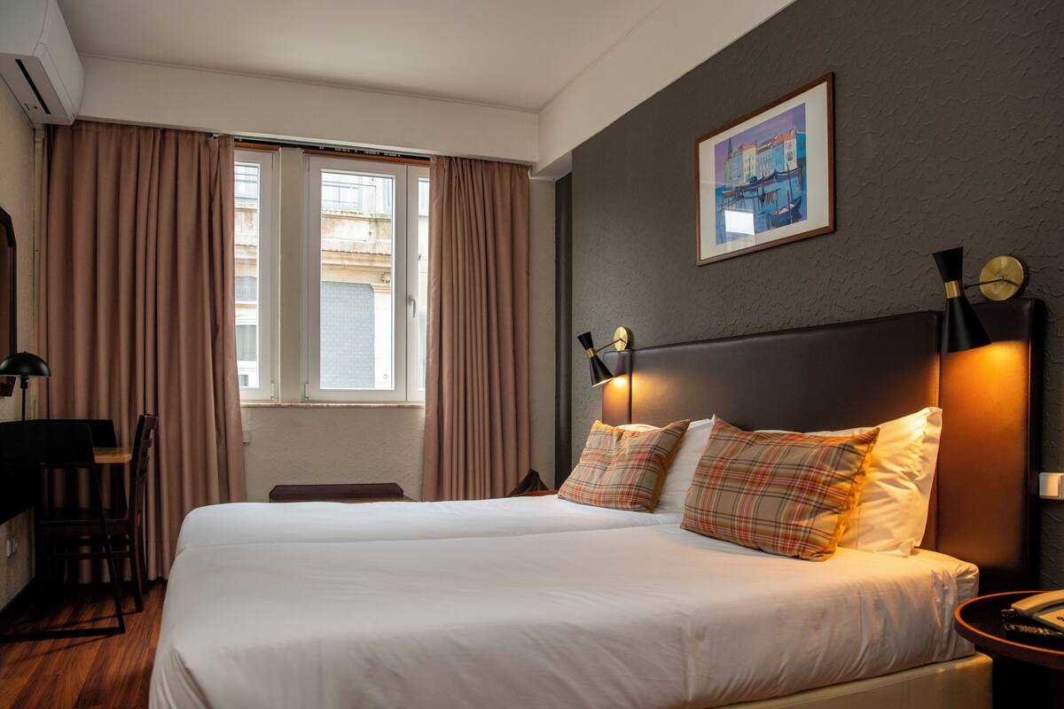 A hotel room with a large bed, a desk with a chair, a window with curtains, and a wall-mounted clock.