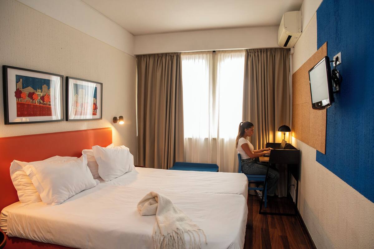 A hotel room with a bed, a desk, a chair, a TV, and two framed pictures on the wall.