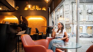 A modern cafe with leather chairs and a glass table, a woman holding a glass, and another woman at the counter. - WOT Porto Soul (Porto)