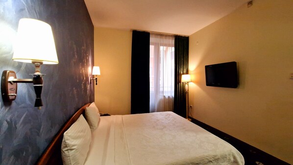 A hotel room with a bed, two wall-mounted lamps, a television, and a window with curtains.