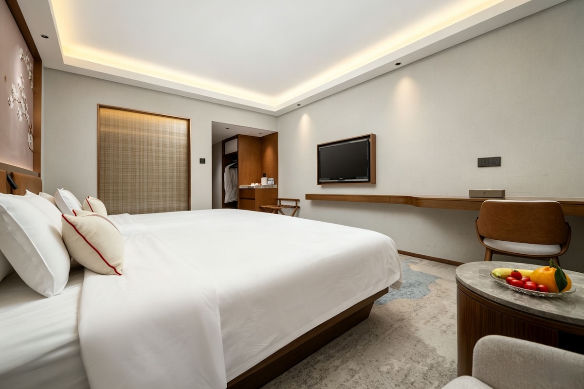 A modern hotel room with a large bed, a TV mounted on the wall, a desk with a chair, and a small table with fruit.