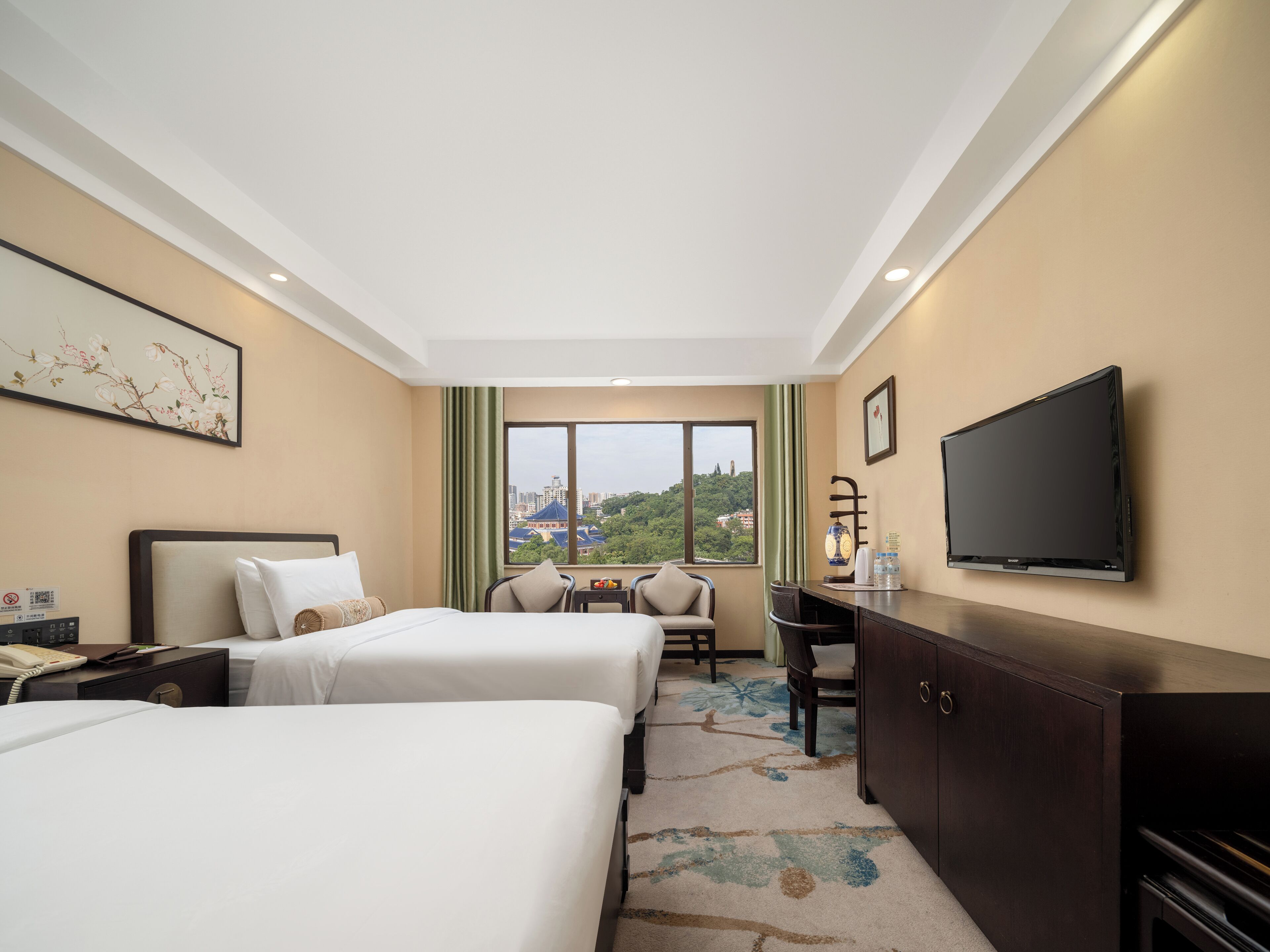 A hotel room with two beds, a desk, a TV, and a view of a cityscape.