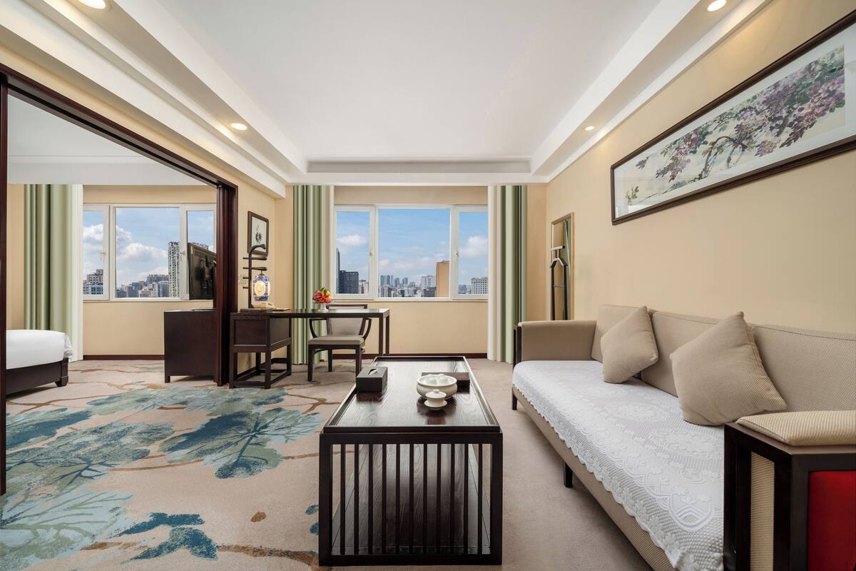 A hotel room with a bed, a sofa, a dining table, and a view of the cityscape through large windows.