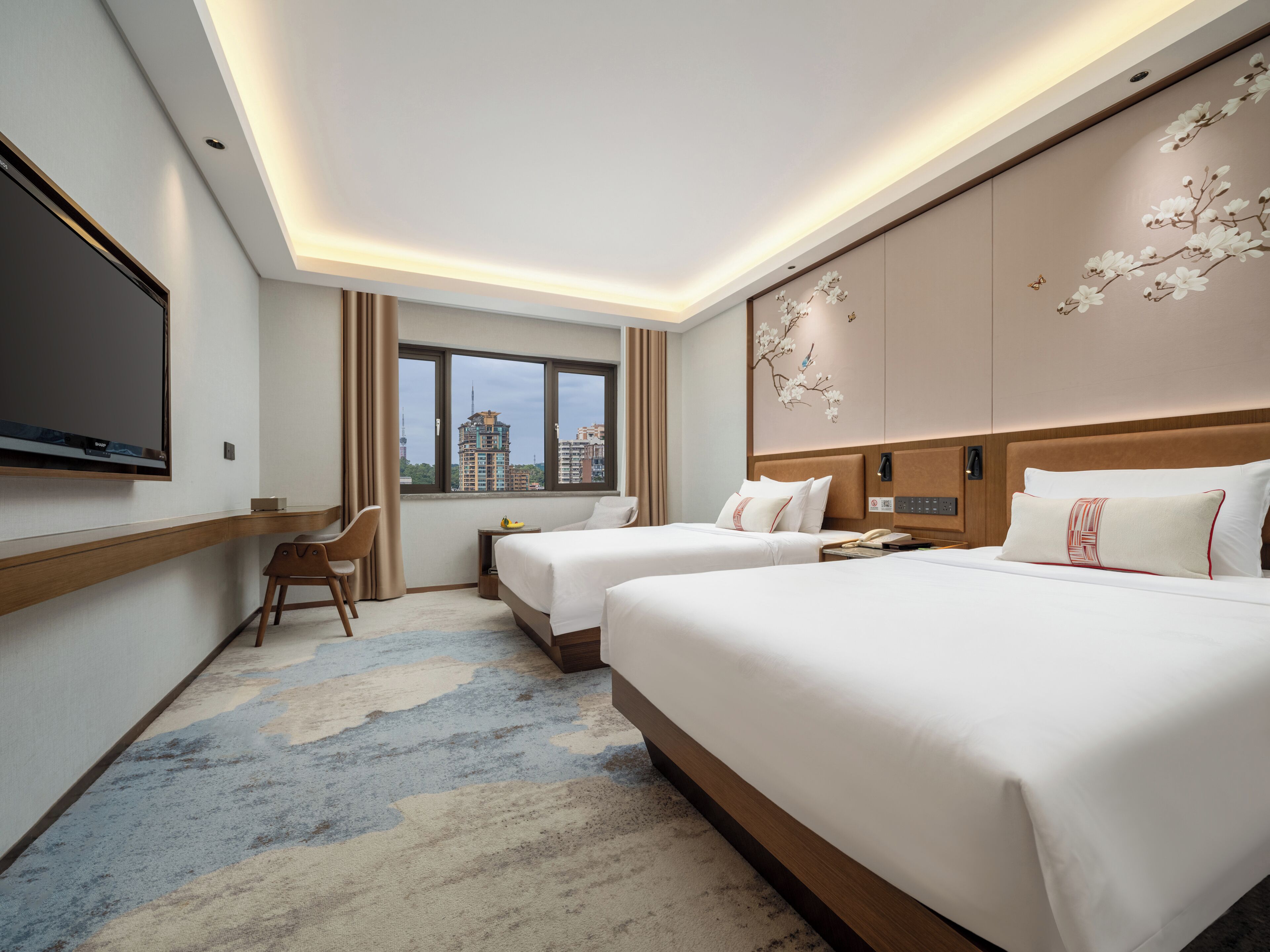 A hotel room with two beds, a desk, a TV, a window with a city view, and a decorative wall piece.