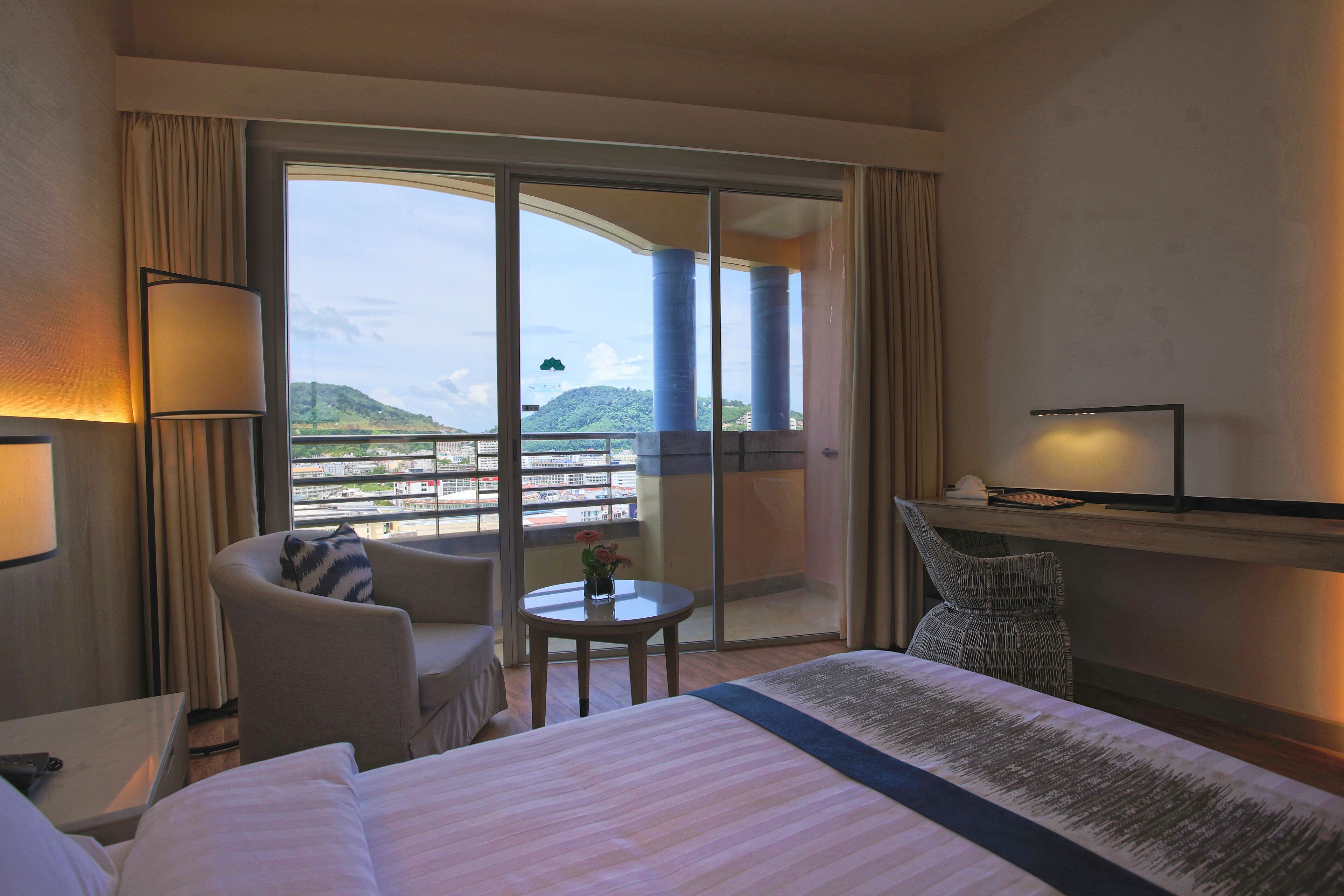 A hotel room with a large bed, a sitting area with a chair and a small table, a TV on a stand, and a balcony with a view of the city and hills.
