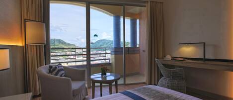 A hotel room with a large bed, a sitting area with a chair and a small table, a TV on a stand, and a balcony with a view of the city and hills.