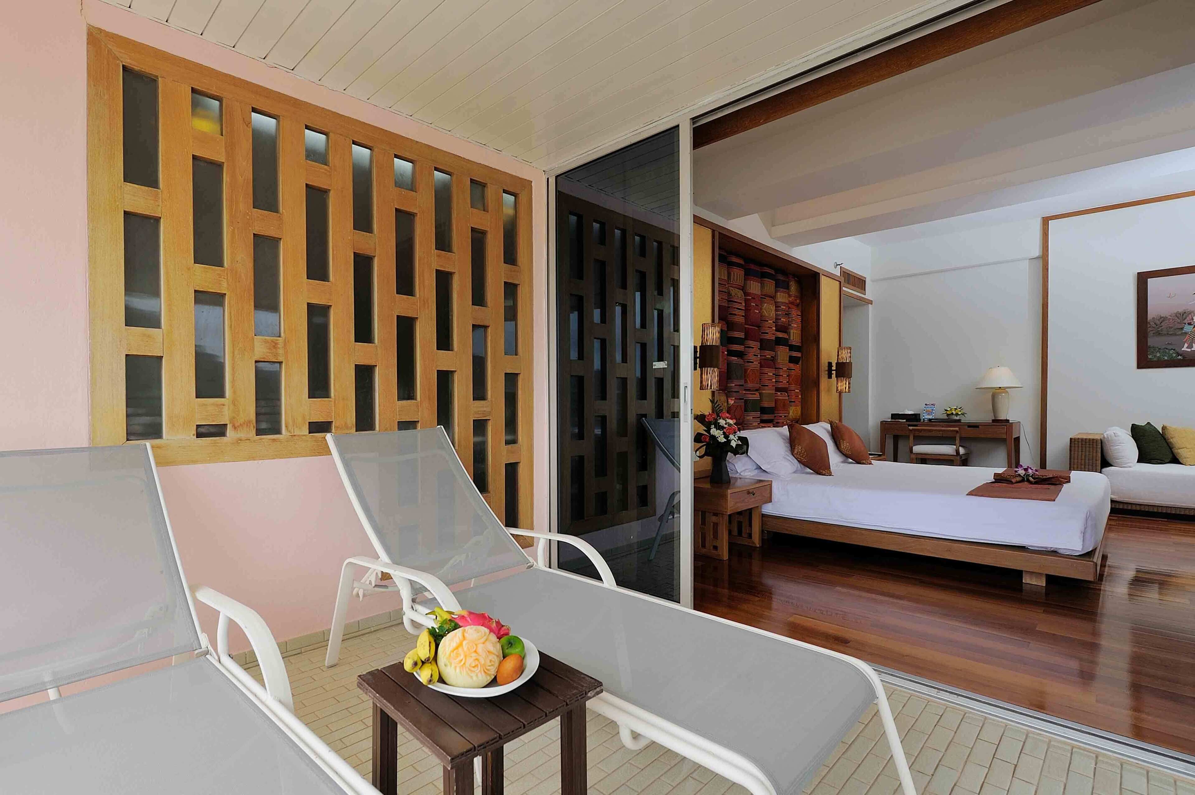 A hotel room with a balcony, a bed, a desk, and a dining table with fruit.