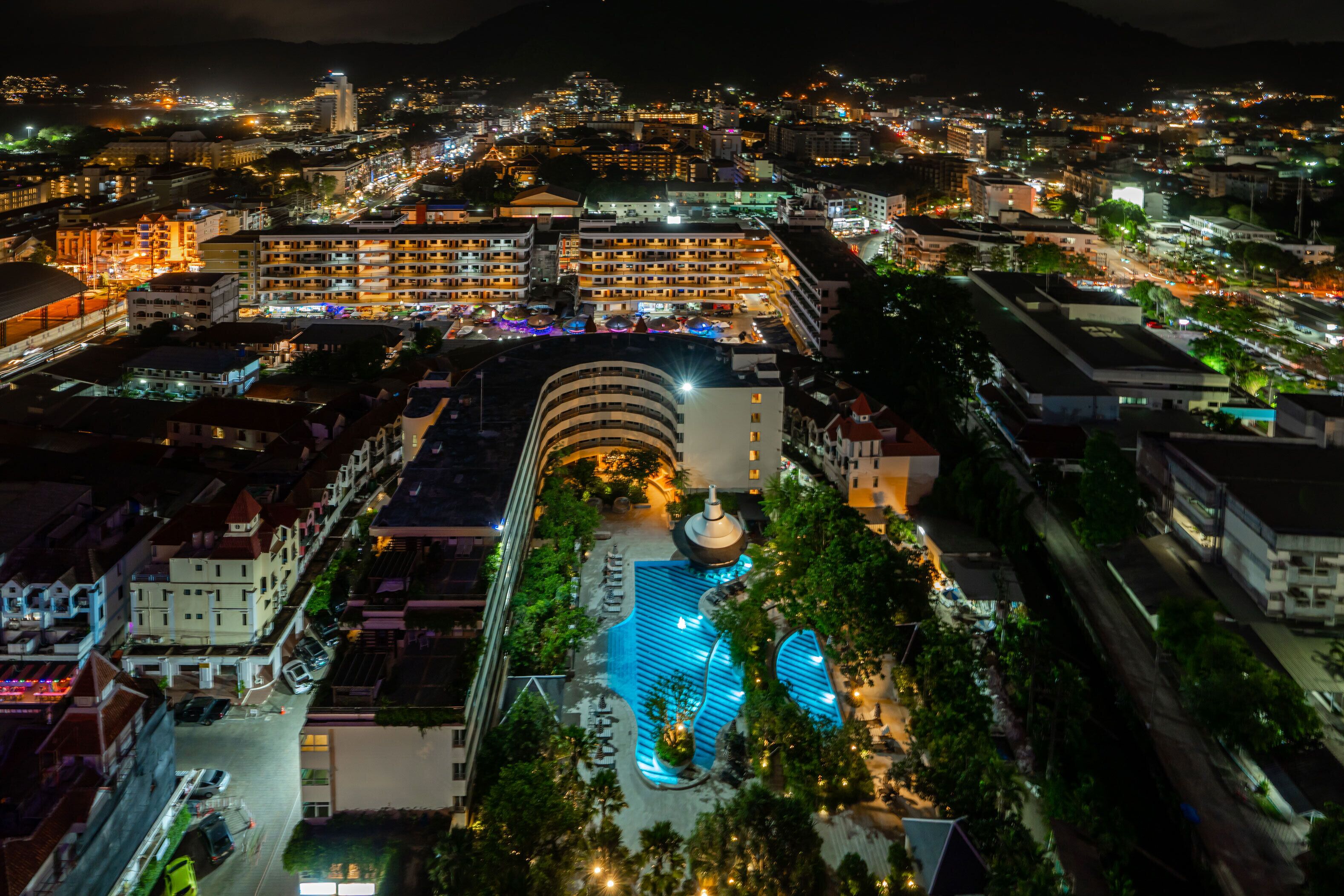 Vista aérea de un hotel con piscina, rodeado de otros edificios y montañas al fondo.