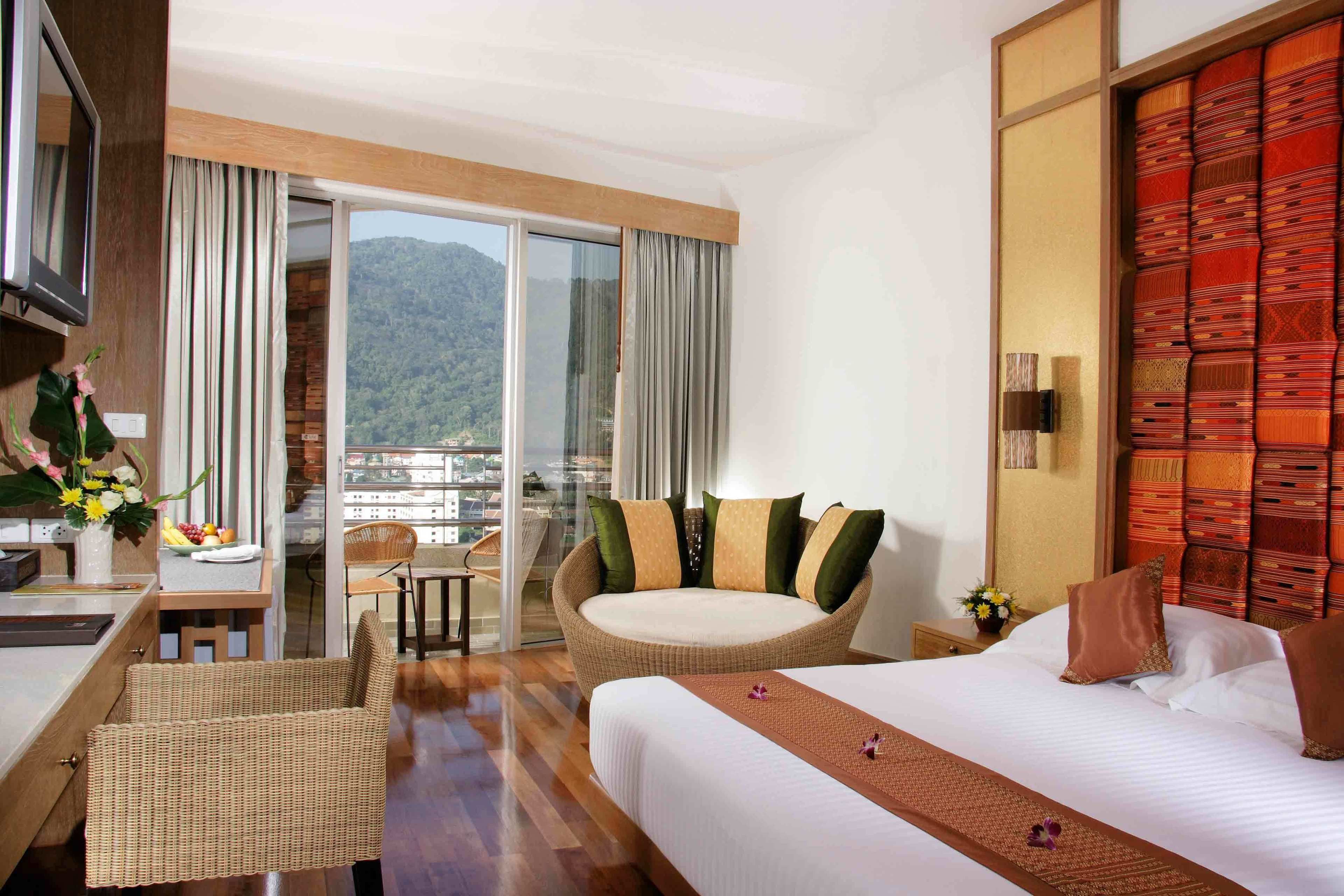 A hotel room with a bed, a desk, a chair, a sofa, and a view of mountains through a sliding glass door.