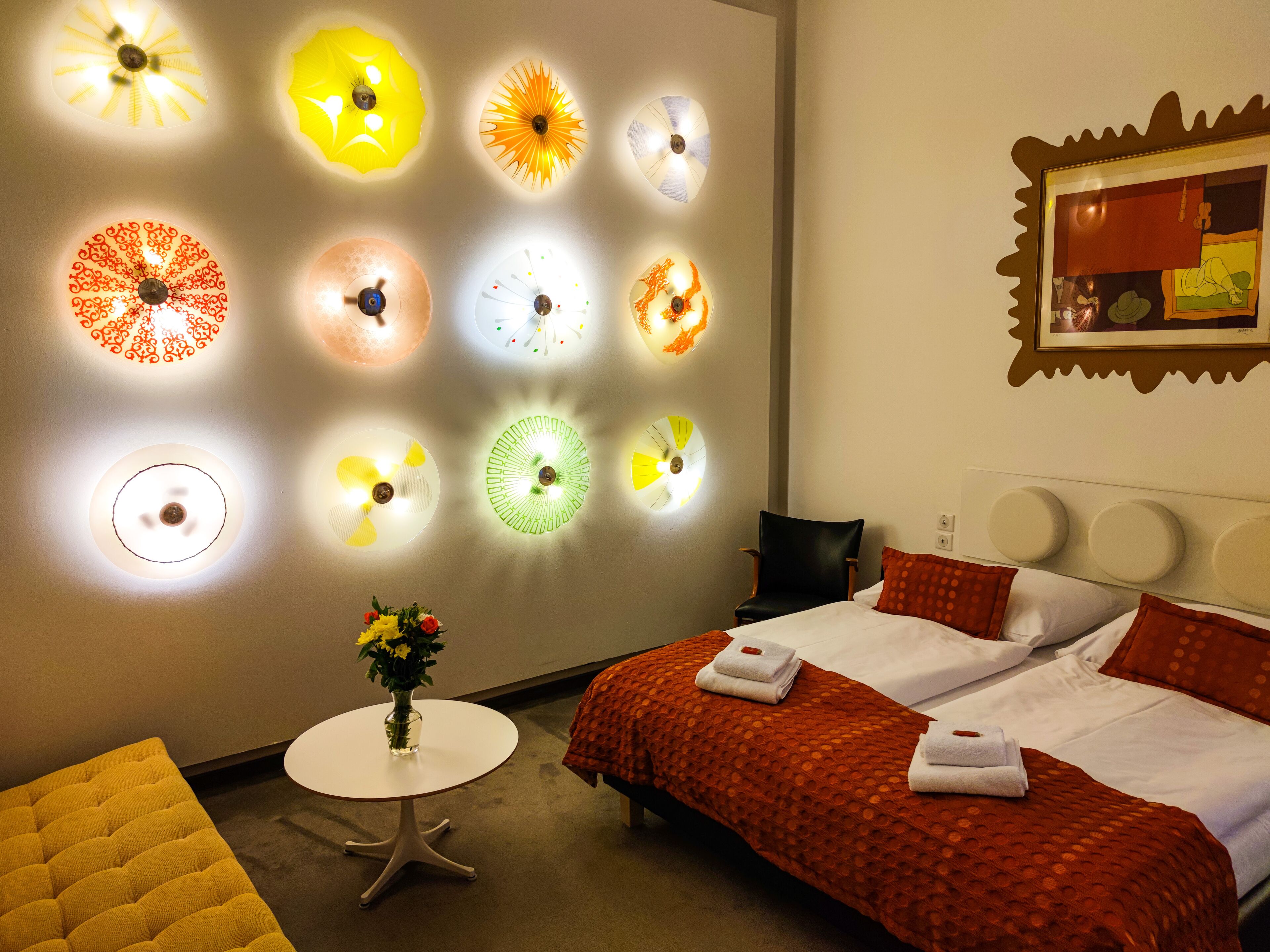 A hotel room with a bed, a round table with a vase of flowers, and a wall with multiple ceiling lights.