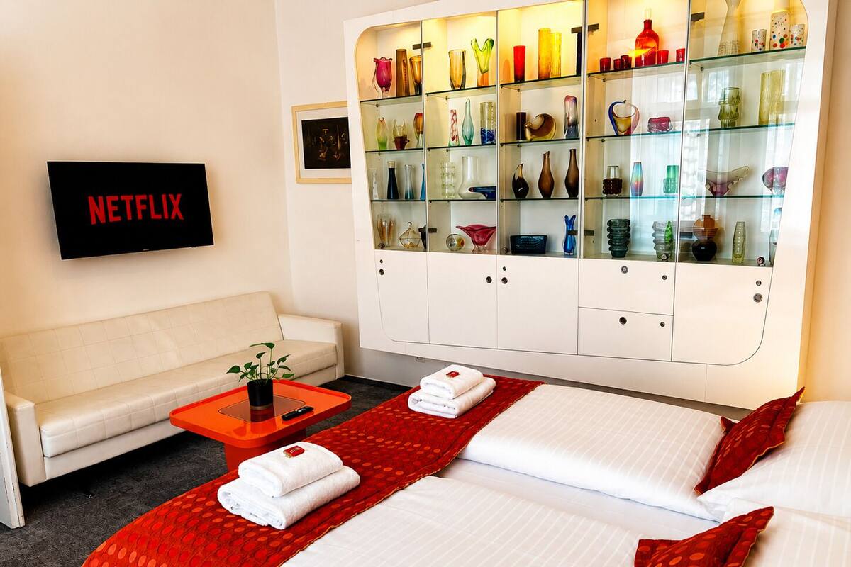 A hotel room with a sofa, two beds, a glass cabinet with decorative items, a TV, and a red polka dot area rug.