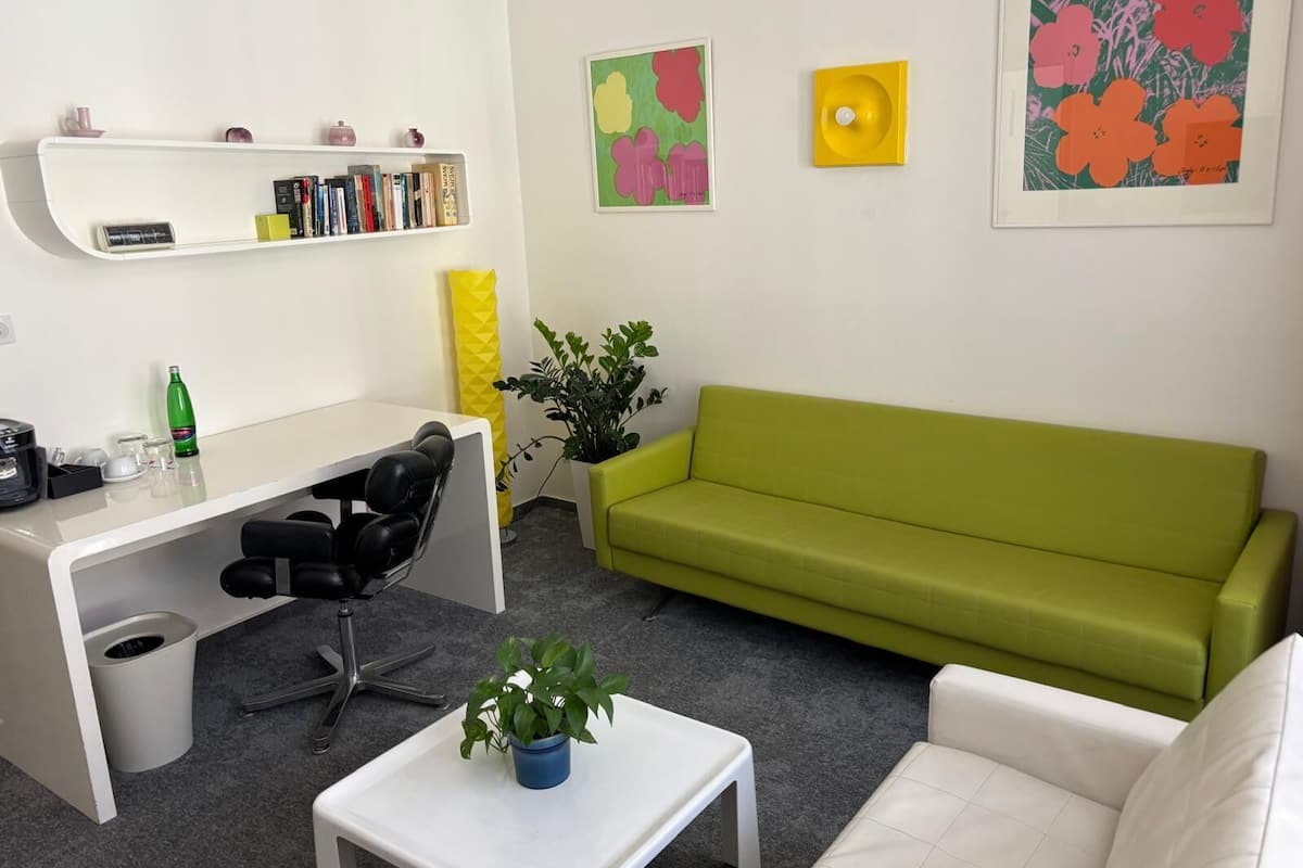A modern living room with a green sofa, a white coffee table with a plant, a desk with a computer and books, and a yellow decorative item on the wall.