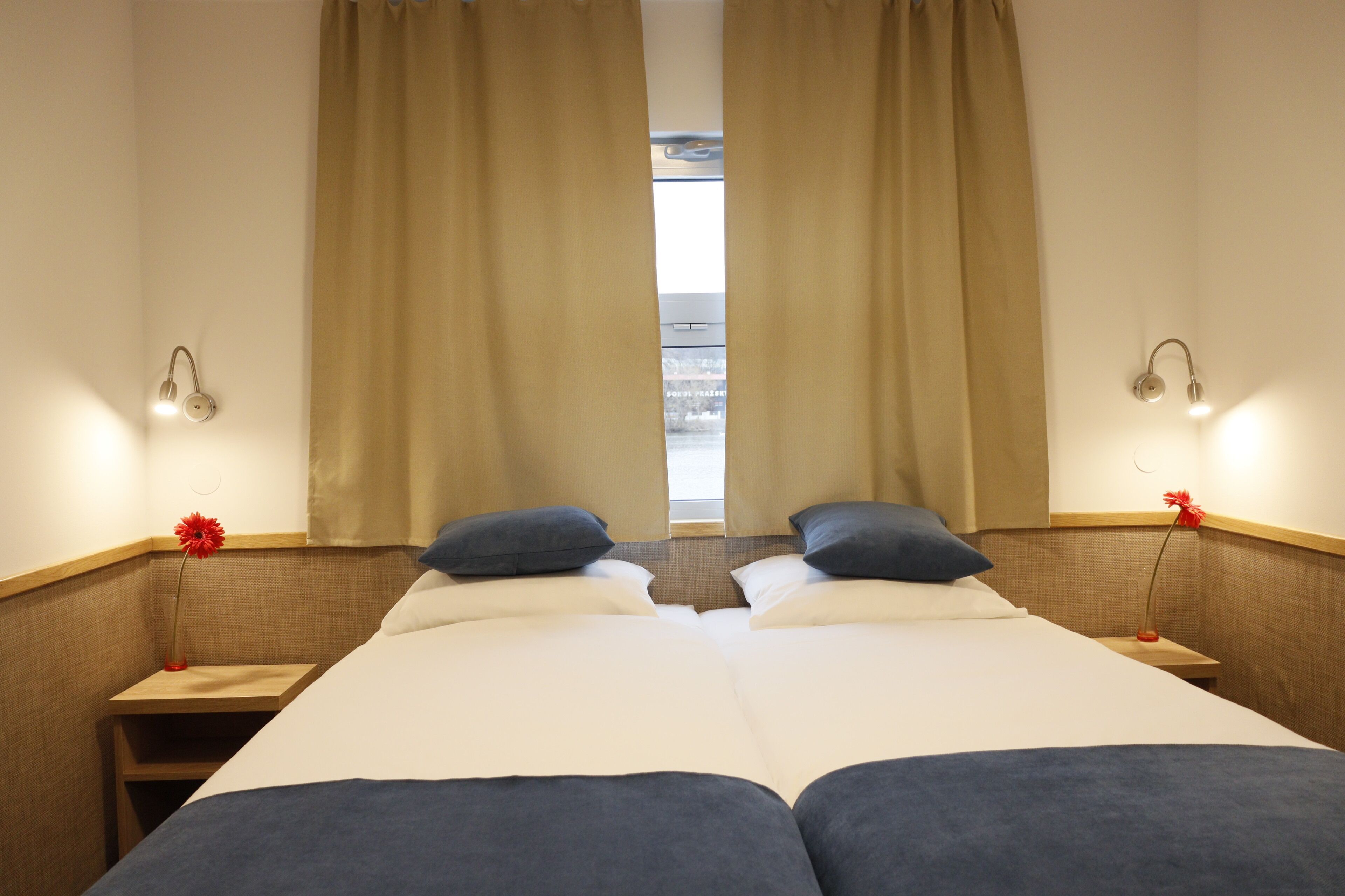 A hotel room with two beds, a nightstand with a lamp, and a view through the curtains.