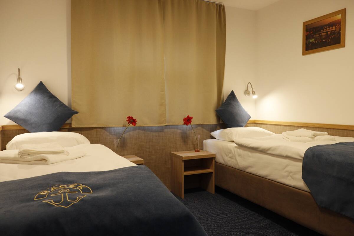A hotel room with two beds, a nightstand, a small table, a window with curtains, and a wall-mounted light.