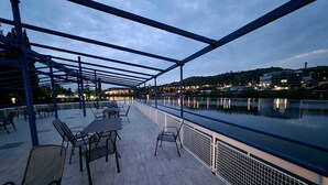 An outdoor terrace with tables and chairs, overlooking a body of water and a cityscape at dusk. - Botel Racek (Prague)