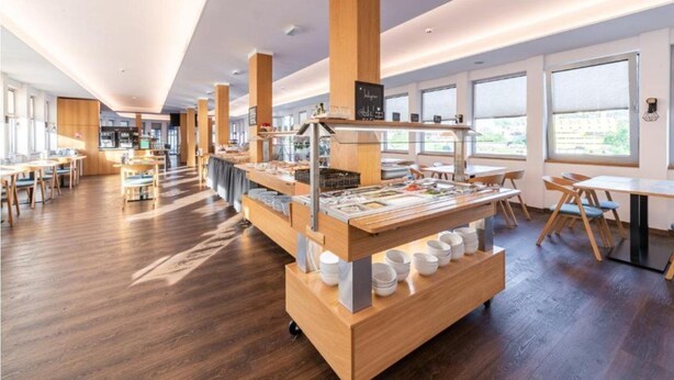 A modern cafe with wooden furniture, a central serving counter, and large windows. - Botel Racek (Prague)