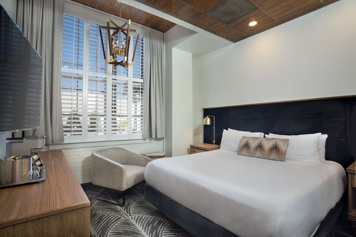 A hotel room with a large bed, a TV, a bathtub, and a window with blinds.