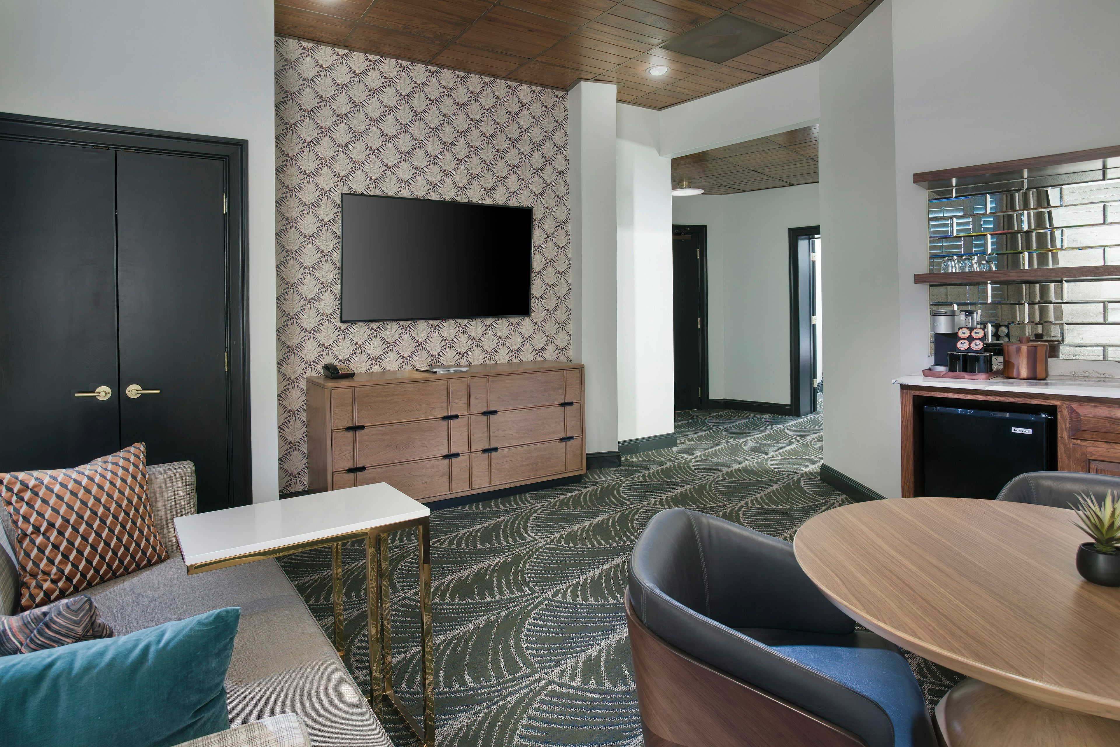 A modern interior with a flat-screen TV mounted on a patterned wall, a wooden cabinet, a dining table with chairs, and a sofa with patterned cushions.