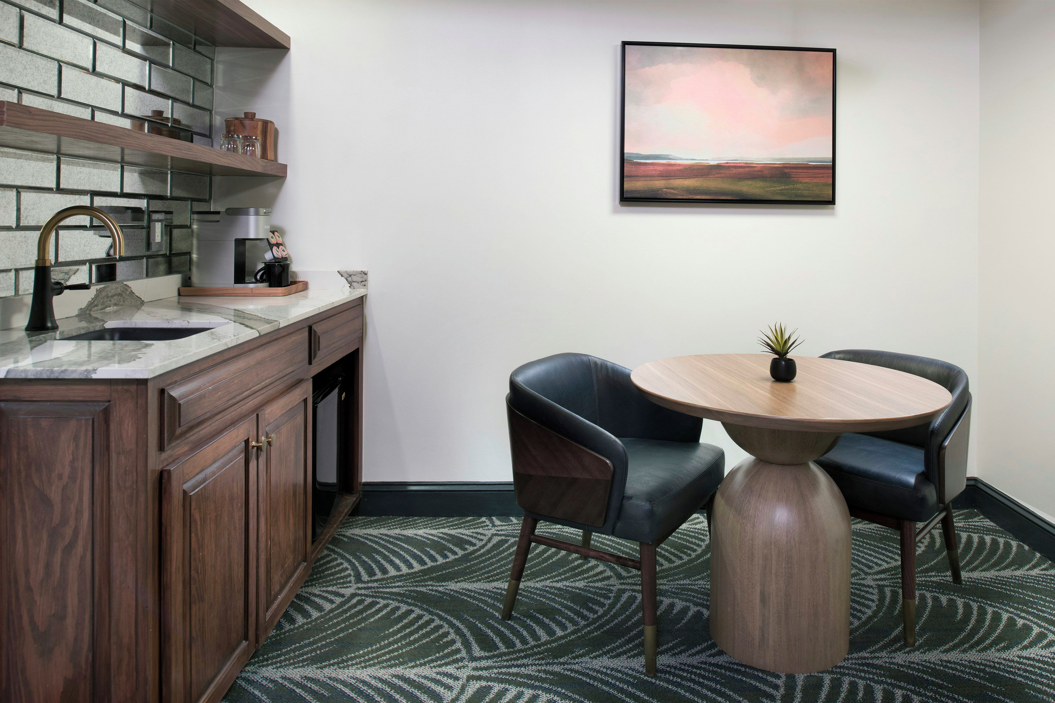 Une cuisine moderne équipée d’un four encastré, d’une machine à café et d’un évier. On y trouve un coin repas avec une table ronde et deux chaises. Un tableau est accroché au mur, au-dessus de la table.