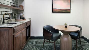 Cozinha moderna com forno embutido, máquina de café e pia. Possui uma área de jantar com mesa redonda e duas cadeiras. Um quadro está pendurado na parede acima da mesa.