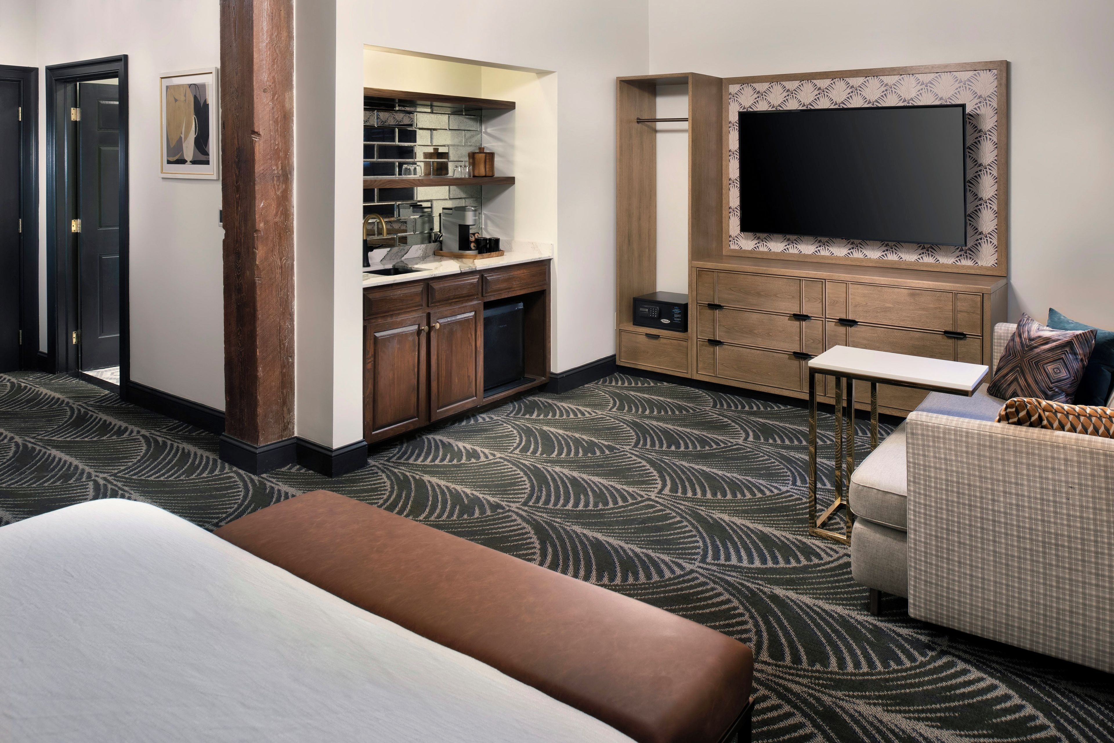 A hotel room with a bed, a sofa, a TV, a kitchenette, and a patterned carpet.