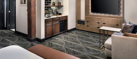 A hotel room with a bed, a sofa, a TV, a kitchenette, and a patterned carpet.