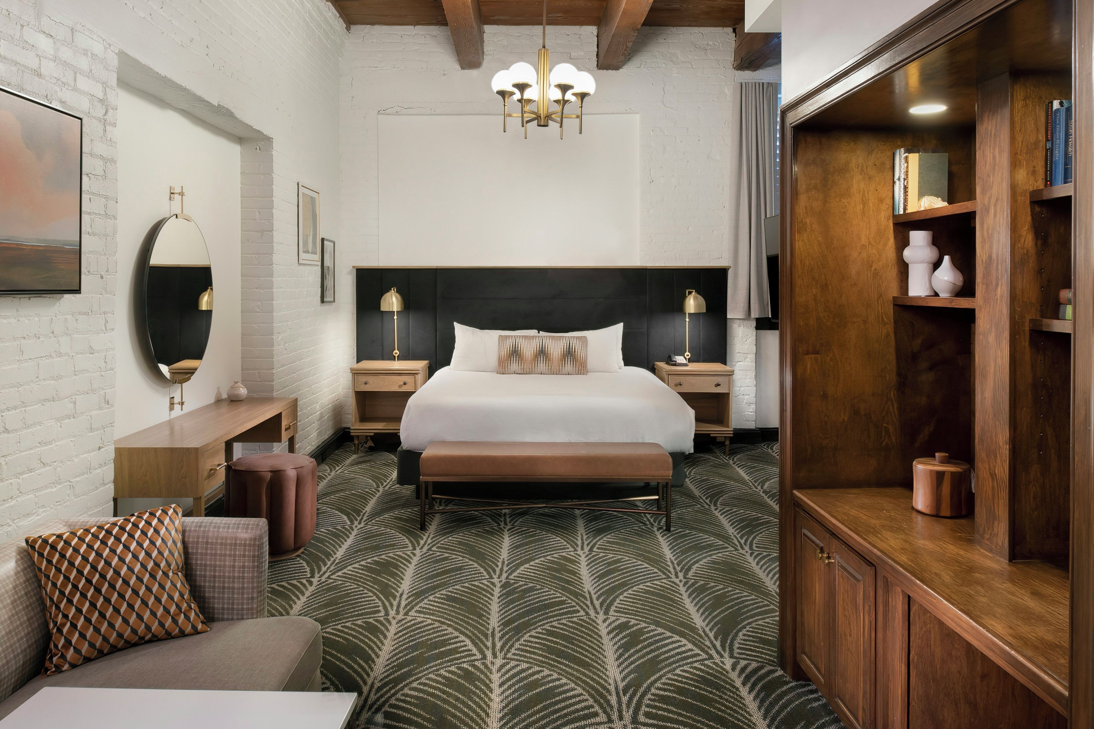 Ein modernes Hotelzimmer mit einem großen Bett, einer hölzernen Bank, einem runden Beistelltisch, einem Spiegel und einem Bücherregal.