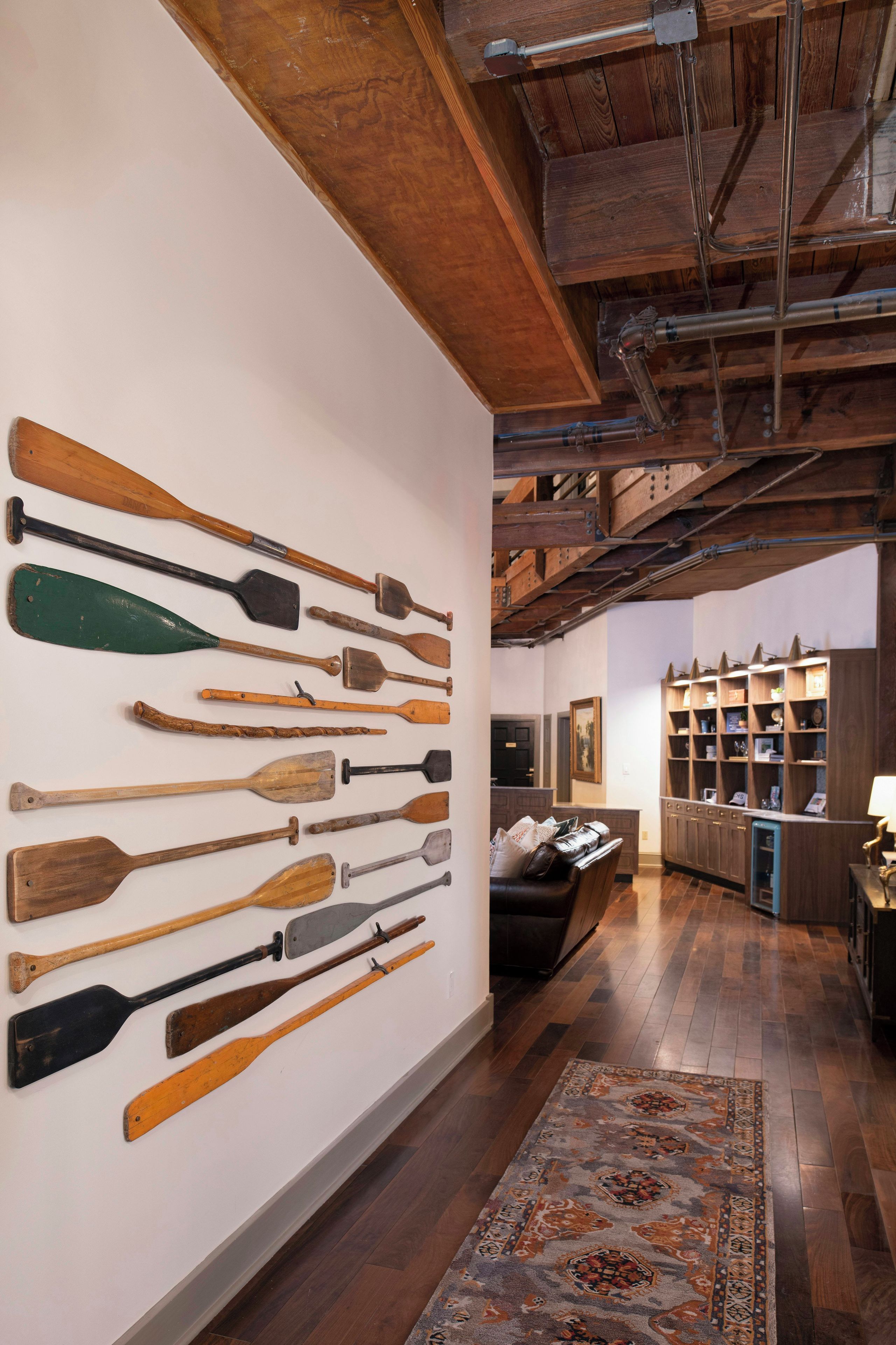 A hallway with a wall displaying various wooden paddles, a patterned rug, and a wooden ceiling.