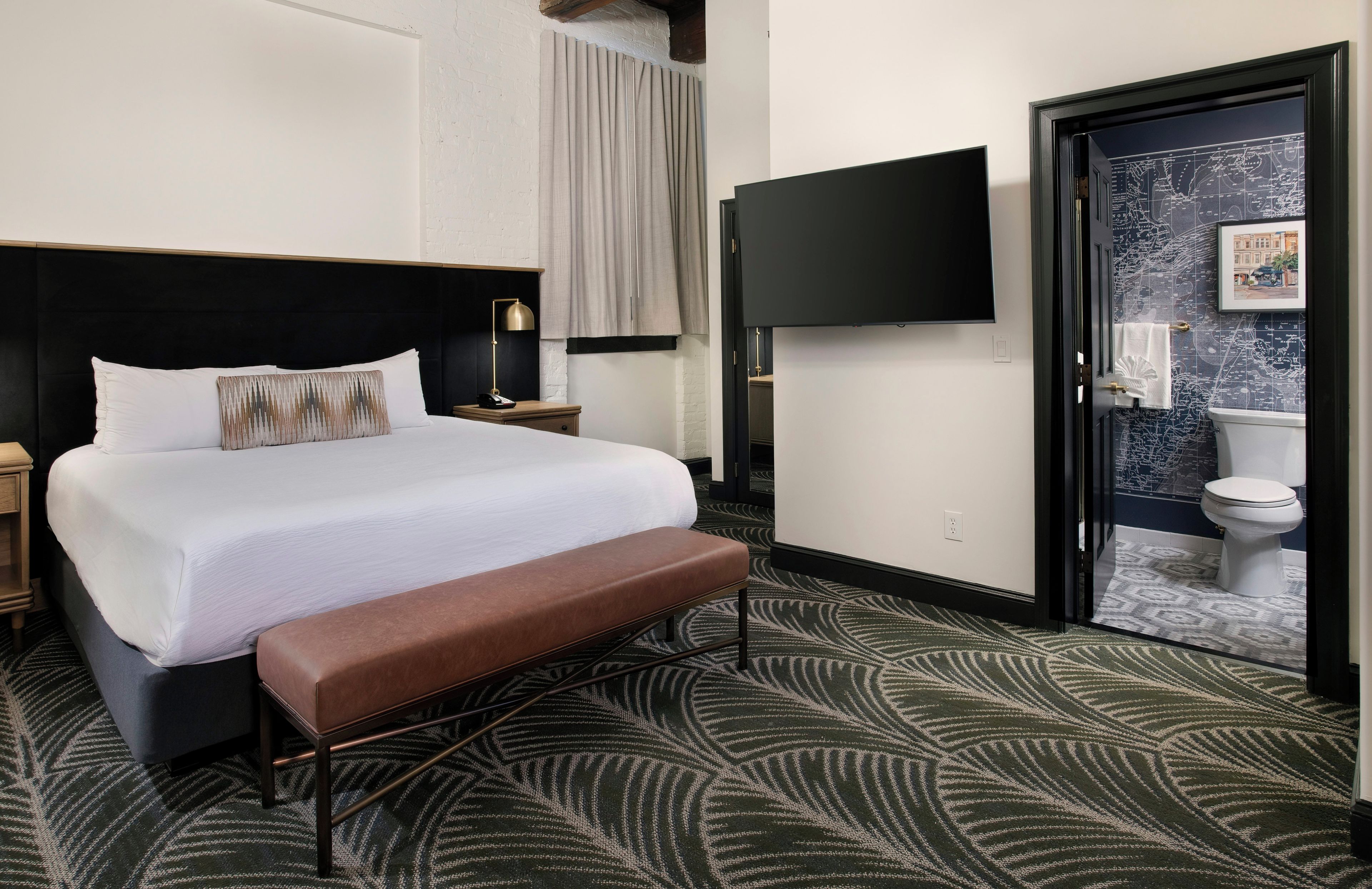 Habitación de hotel con cama, un banco, un televisor y un baño visible a través de una puerta abierta.