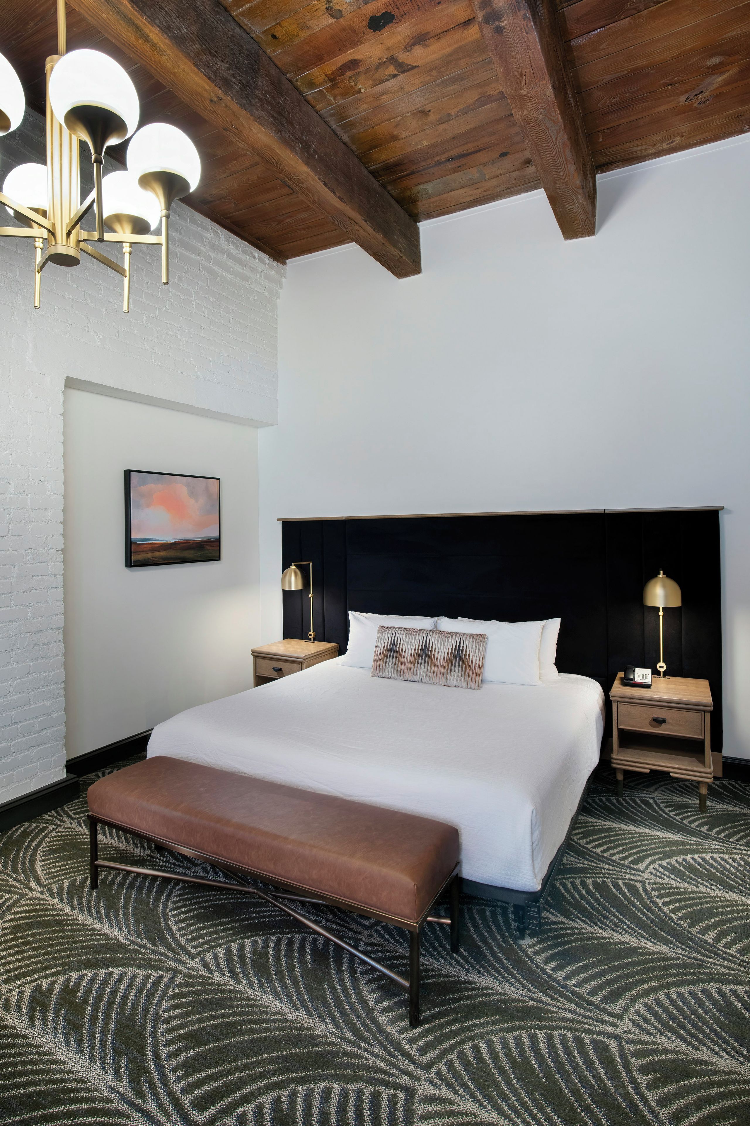 A hotel room with a large bed, a bench, a headboard, bedside tables, and a ceiling light fixture.