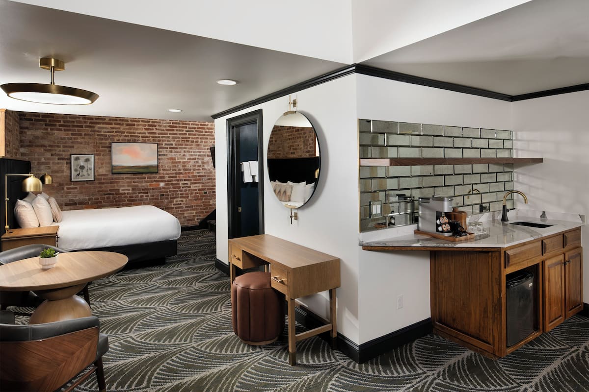 A modern hotel room with a bed, desk, and kitchenette.