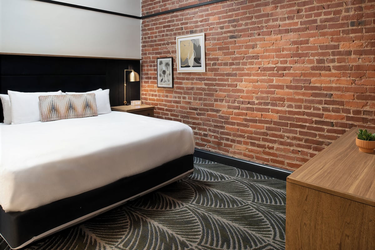 A hotel room with a brick wall, a bed with white bedding, a nightstand with a lamp, and a small plant on the desk.
