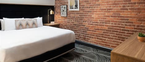 A hotel room with a brick wall, a bed with white bedding, a nightstand with a lamp, and a small plant on the desk.