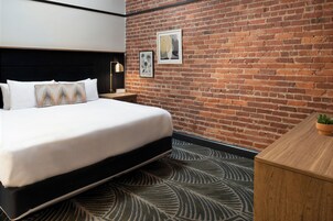 A hotel room with a brick wall, a bed with white bedding, a nightstand with a lamp, and a small plant on the desk.