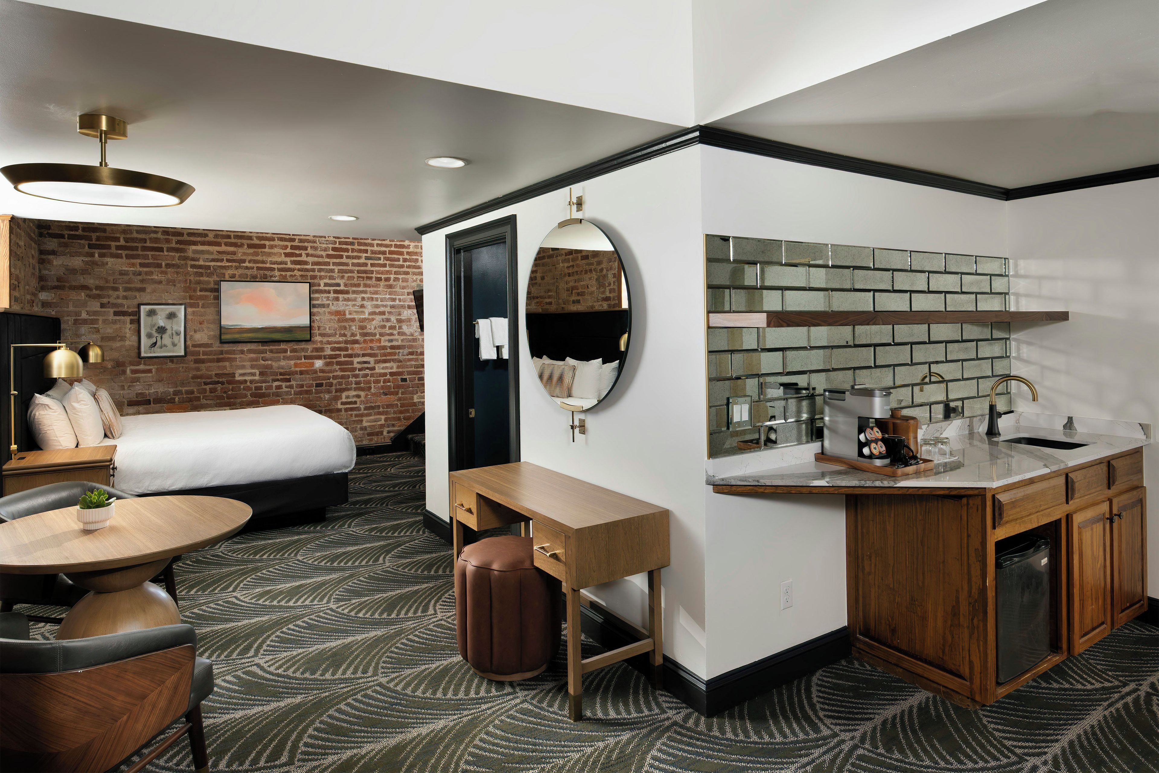 A hotel room with a bed, a desk, a round table, a mirror, a brick wall, and a kitchen area with a sink and coffee maker.