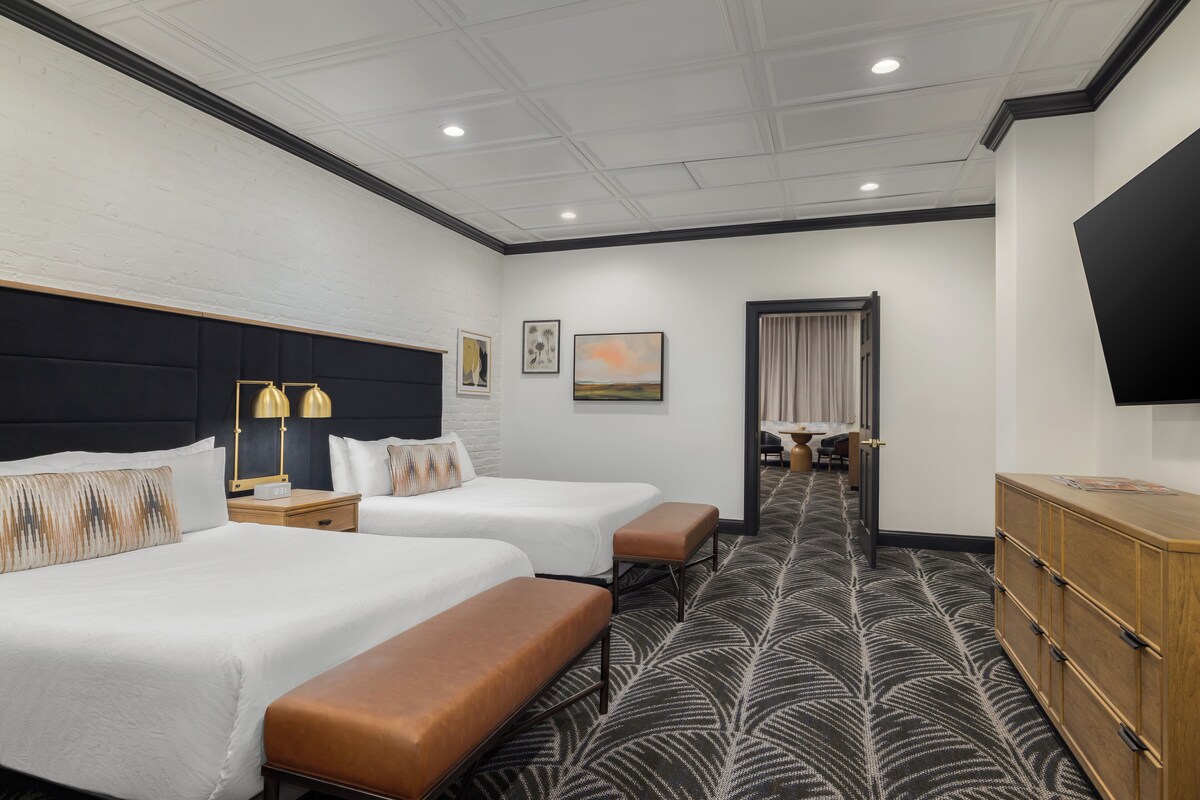 A hotel room with two beds, a desk, a TV, and a view of the hallway.