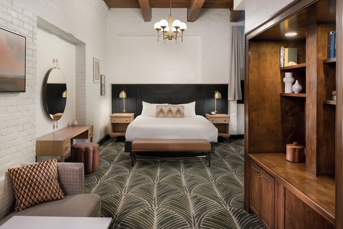 A hotel room with a large bed, a wooden bench, a round mirror, and a wooden cabinet with shelves.
