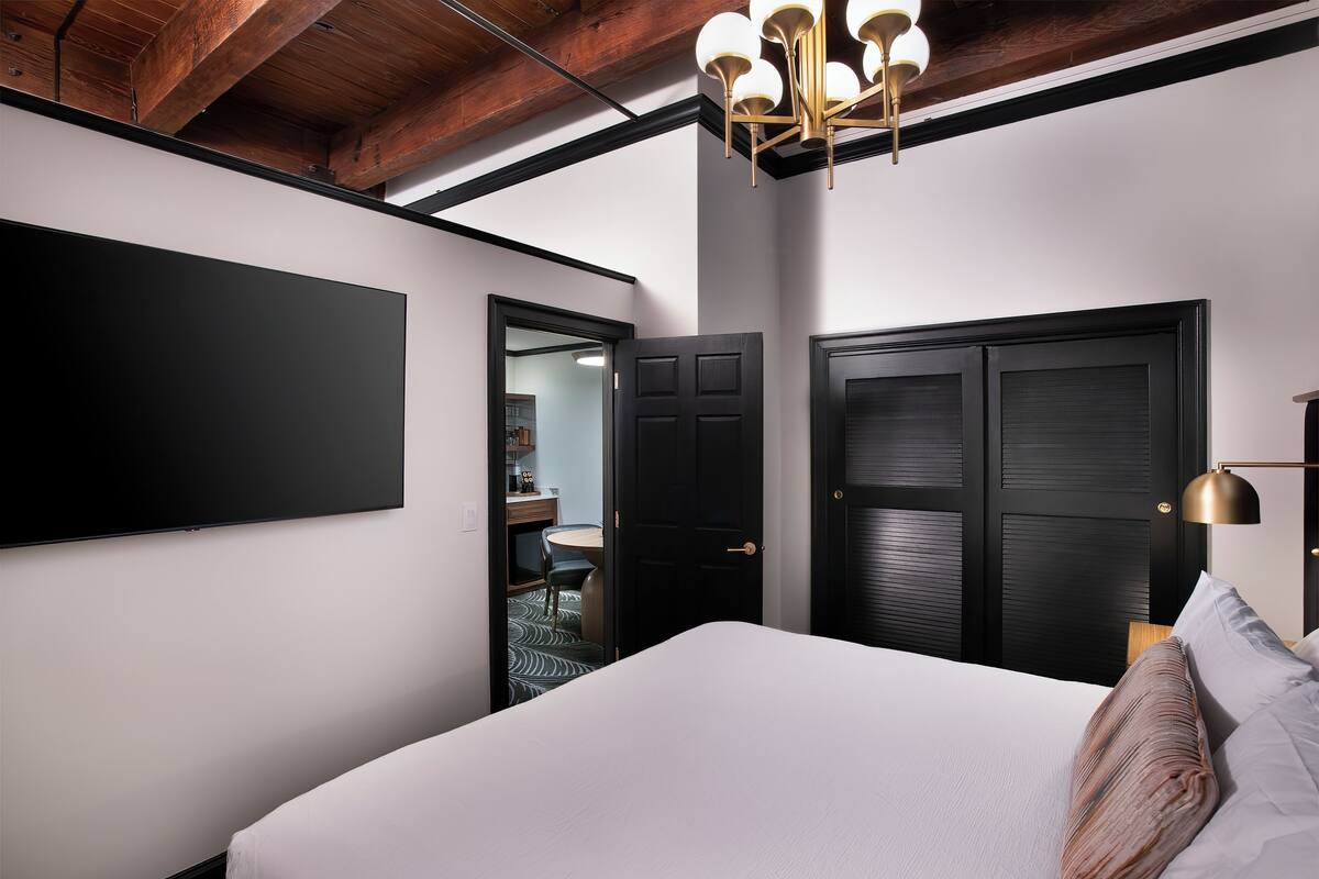 A modern hotel room with a large bed, a flat-screen TV mounted on the wall, and a dark wooden door.