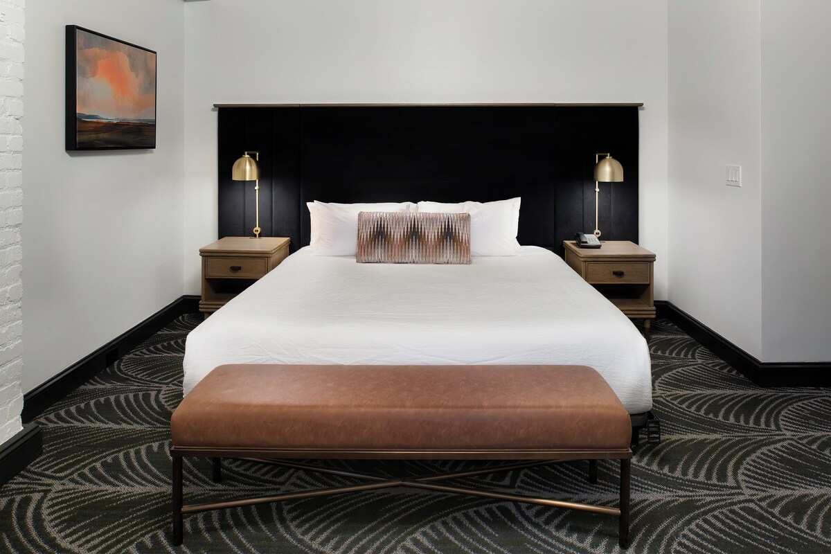 A hotel room with a large bed, two bedside tables with lamps, a bench, and a patterned carpet.