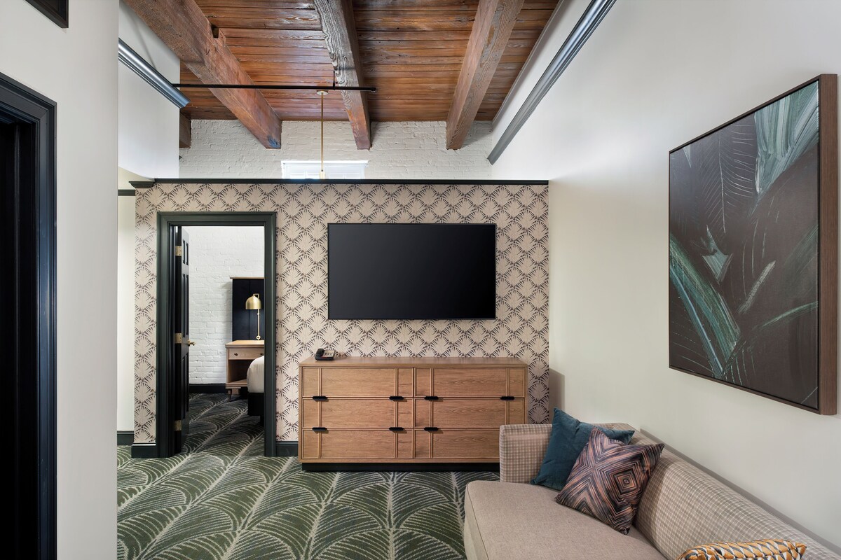A modern hotel room with a large flat-screen TV mounted on the wall, a wooden cabinet, a patterned carpet, and a sofa with decorative pillows.
