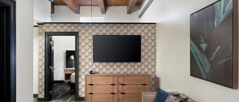 A modern hotel room with a large flat-screen TV mounted on the wall, a wooden cabinet, a patterned carpet, and a sofa with decorative pillows.