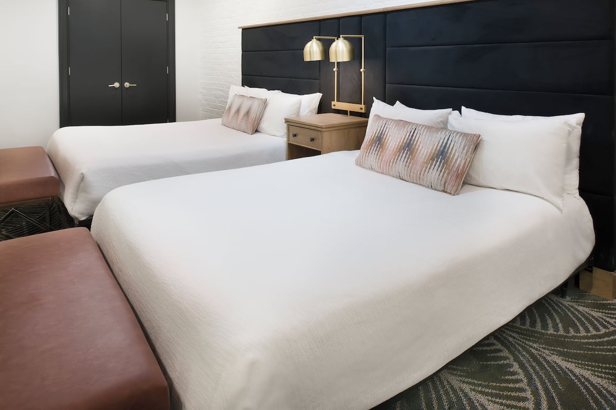 A hotel room with two beds, a nightstand, a lamp, and a bench.