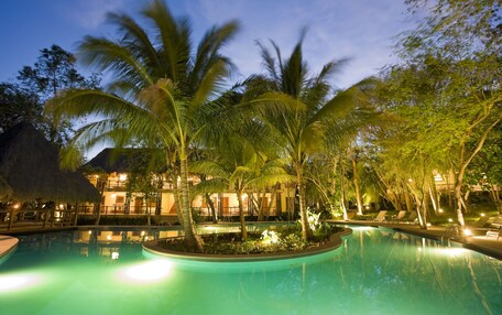 A swimming pool surrounded by palm trees and a building with a thatched roof.