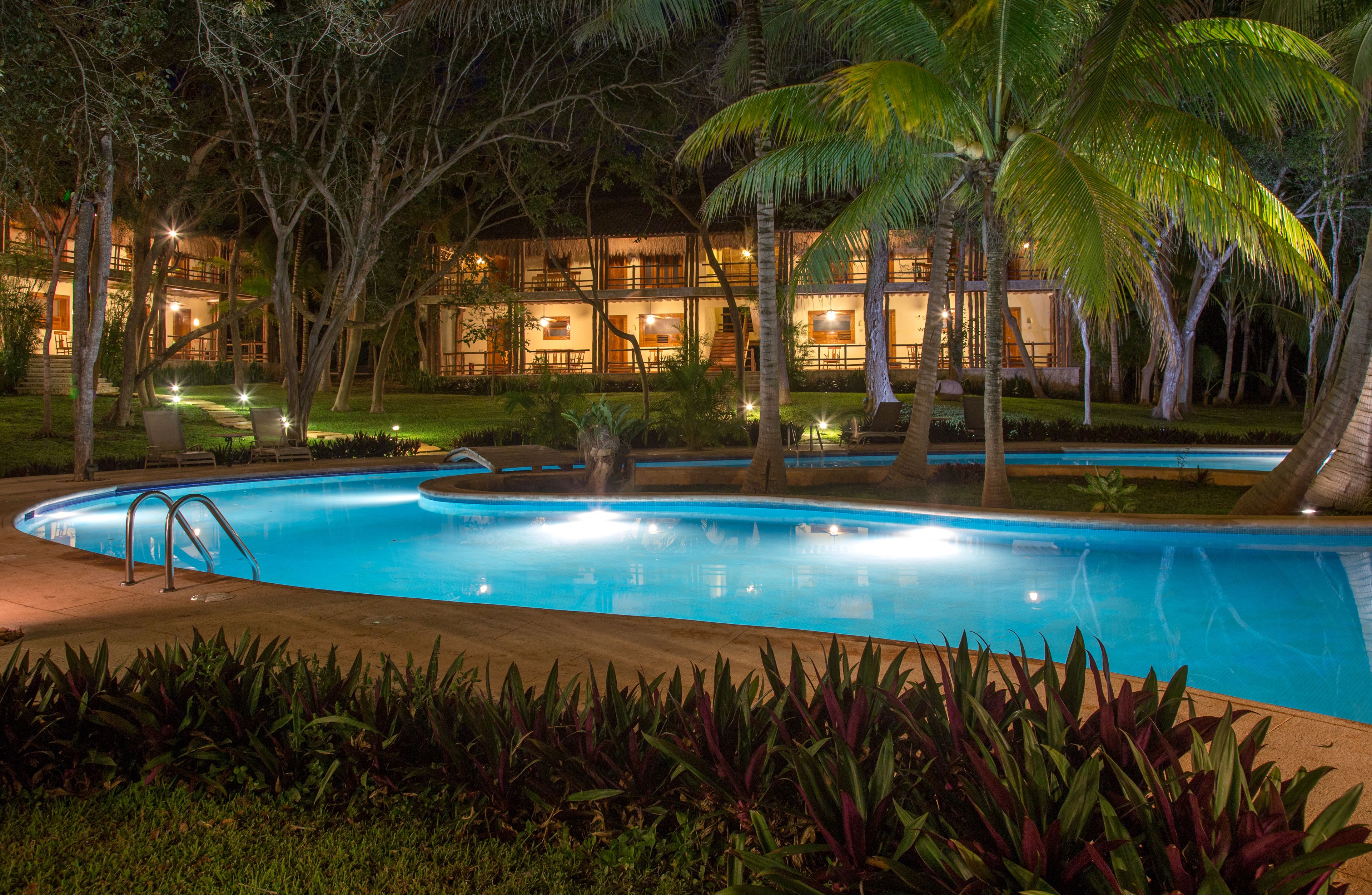 A swimming pool surrounded by tropical vegetation and a building with multiple windows and lights.