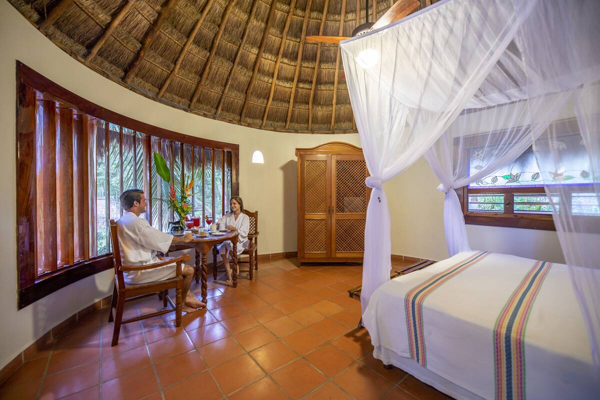 Una habitación con techo de paja, una cama con dosel, un armario de madera y una zona de comedor con mesa y sillas.
