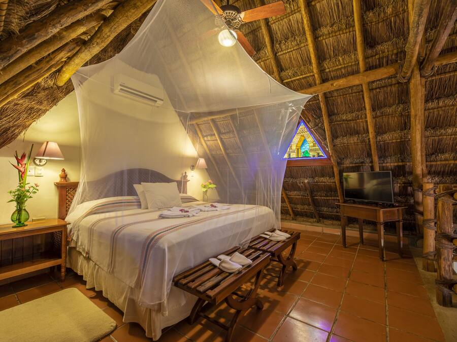Un dormitorio con cama, mesitas de noche, un banco, televisor y ventana con diseño de vitral.