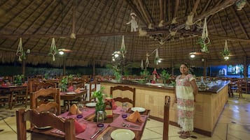 Un restaurante con mesas y sillas de madera, techo de paja, plantas colgantes y una zona de bar central.