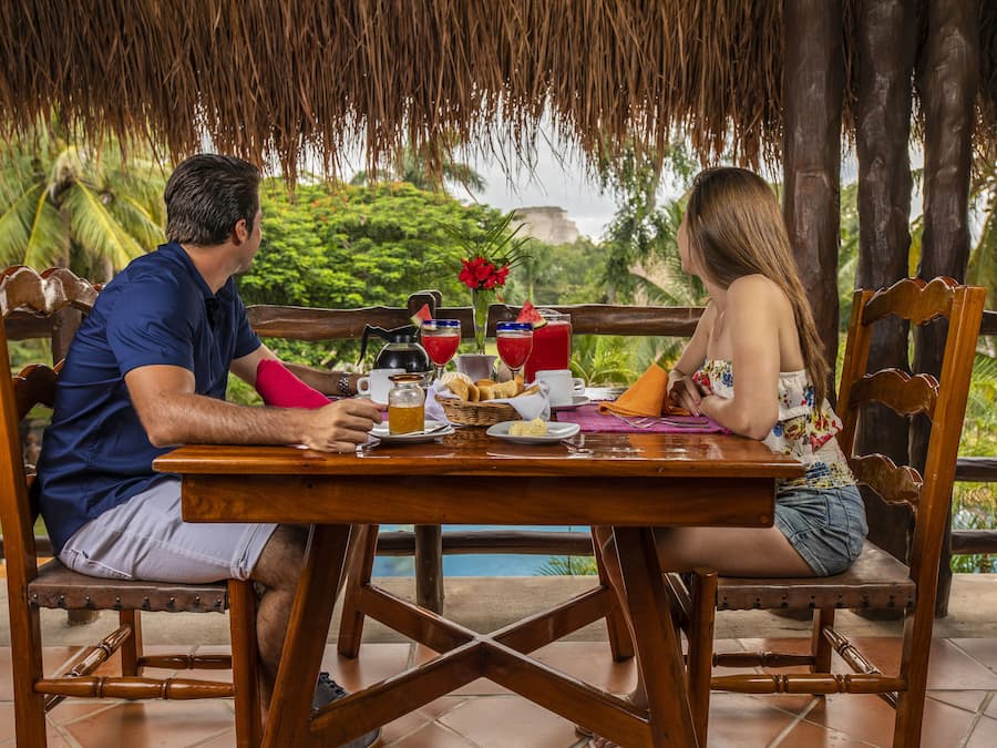 Um casal jantando em uma mesa de madeira, com vista para palmeiras e um telhado de palha.