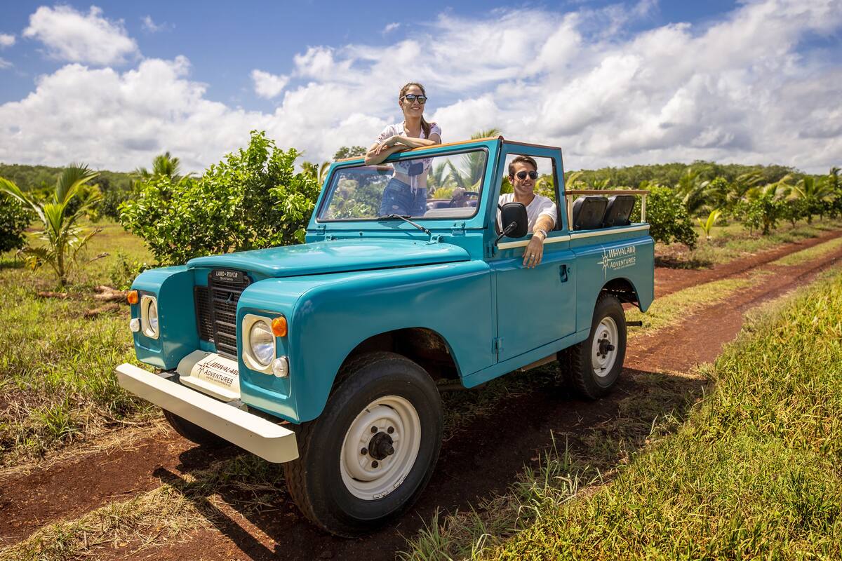 Un Land Rover antiguo color turquesa con dos pasajeros, estacionado en un camino de tierra rodeado de vegetación.